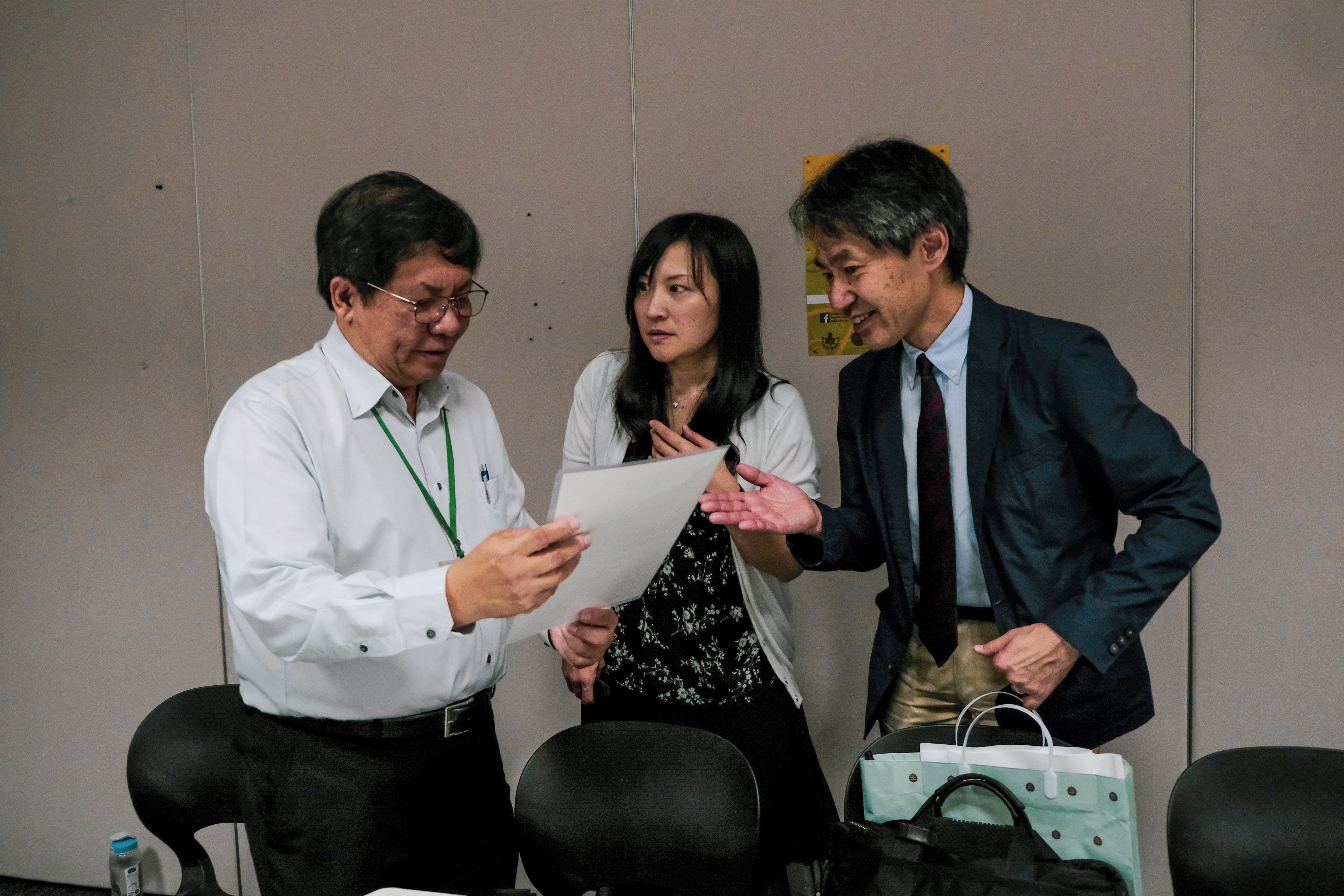 Three people stand together reviewing a document, with a man in a suit pointing at papers held by another man wearing a green lanyard.